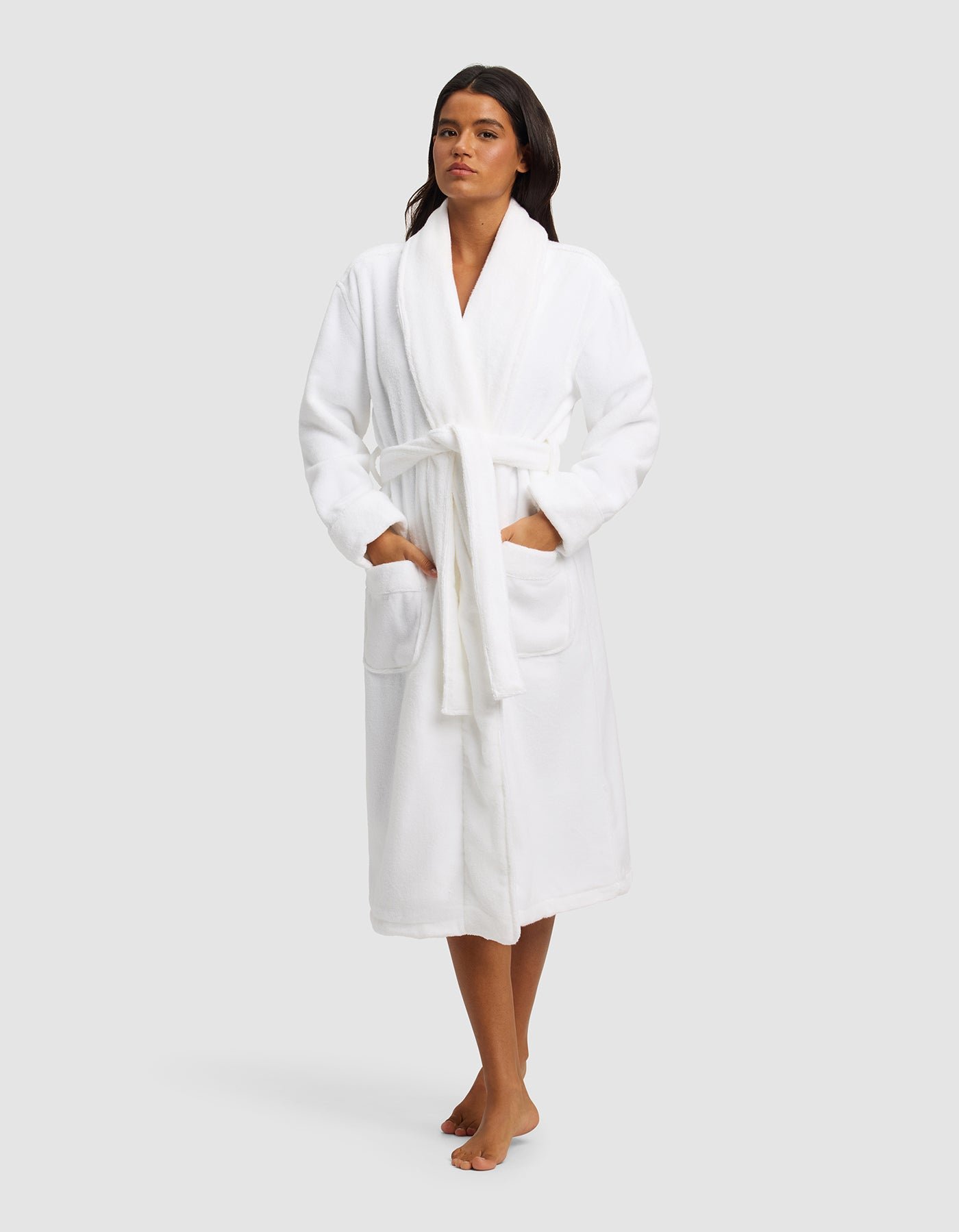A woman with long dark hair poses barefoot against a plain light gray background, wearing the Cozy Earth Luxe Bath Robe in white. The knee-length robe features a tie belt and two front pockets.