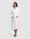 A woman with long dark hair poses barefoot against a plain light gray background, wearing the Cozy Earth Luxe Bath Robe in white. The knee-length robe features a tie belt and two front pockets.|Color:White