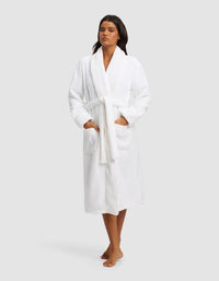A woman with long dark hair poses barefoot against a plain light gray background, wearing the Cozy Earth Luxe Bath Robe in white. The knee-length robe features a tie belt and two front pockets.|Color:White