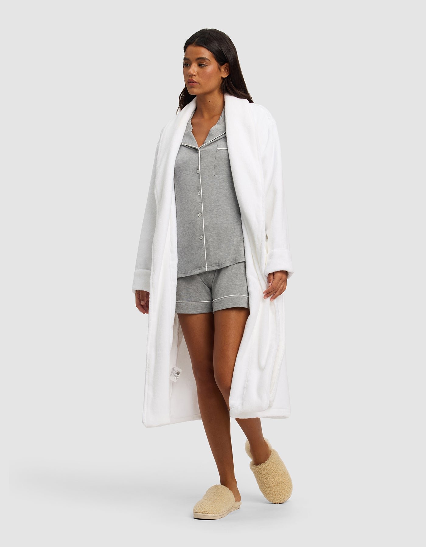 A woman models the Cozy Earth Luxe Bath Robe over gray pajama shorts and matching top, paired with beige fuzzy slippers, against a plain light gray background.