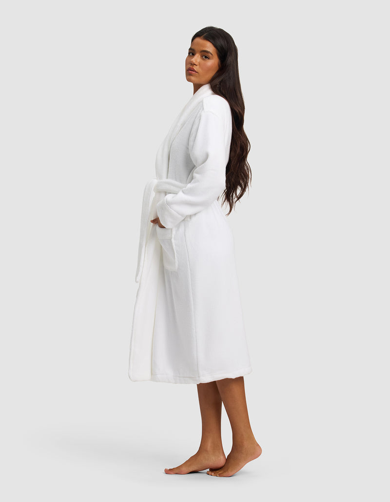 A woman with long dark hair stands barefoot on a white background, wearing the Luxe Bath Robe by Cozy Earth, tied at the waist. She faces slightly to the side with her hands in the robe’s pockets.