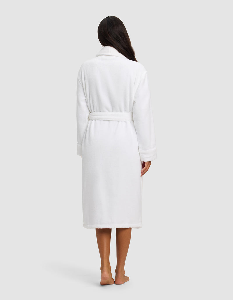A woman with long dark hair stands barefoot facing away, wearing the Cozy Earth Luxe Bath Robe—knee-length, white, and belted—against a plain light background.