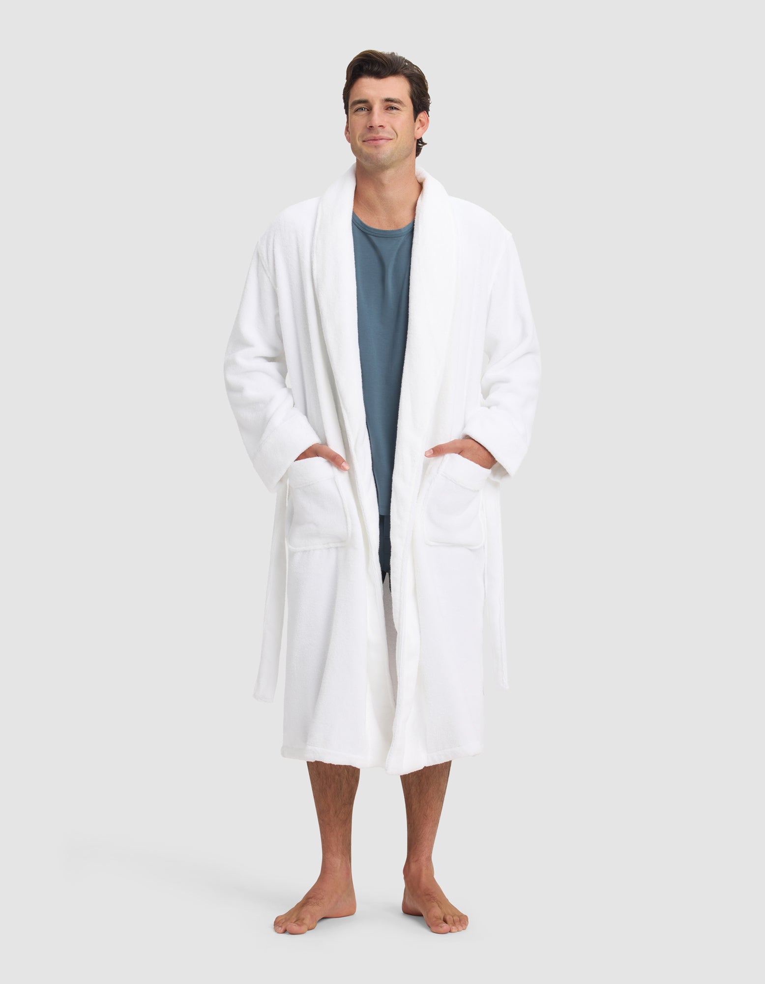 A man stands barefoot against a neutral background, wearing the Cozy Earth Luxe Bath Robe over a blue shirt. His hands are in the robe’s pockets as he looks forward with a slight smile.