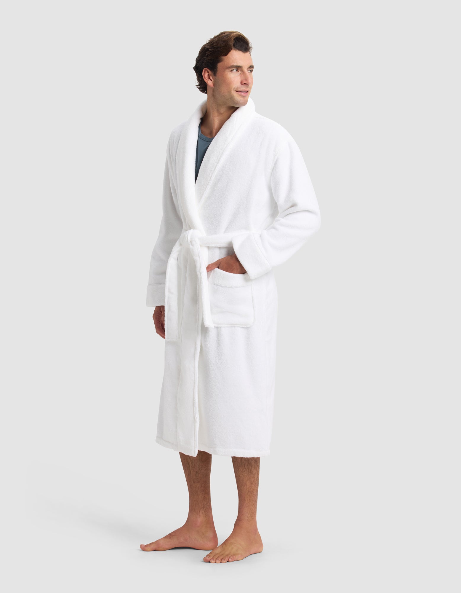 A man stands barefoot on a white background wearing the Cozy Earth Luxe Bath Robe, featuring a belt and large front pockets. He gazes to the side with a relaxed expression.