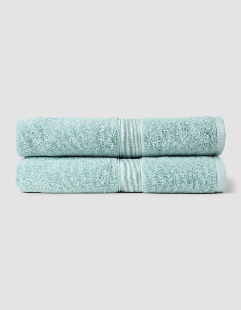 Two Luxe Bath Sheets by Cozy Earth, neatly folded and stacked, feature a light blue color against a plain, light gray background.