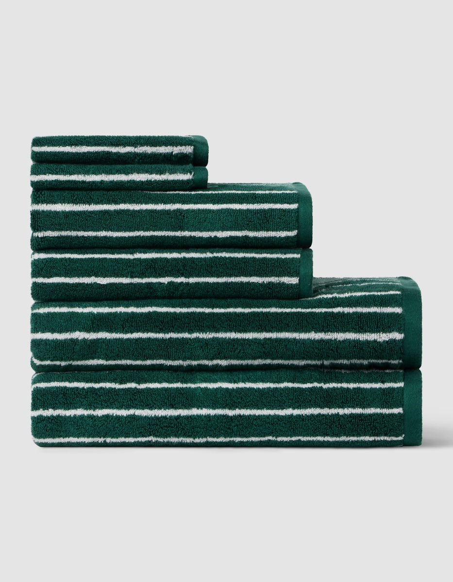 Four Luxe Bath Towel Set towels by Cozy Earth, stacked in ascending order, feature green fabric with horizontal white stripes against a plain light gray background.|Color:Uniform with Seashell Stripe