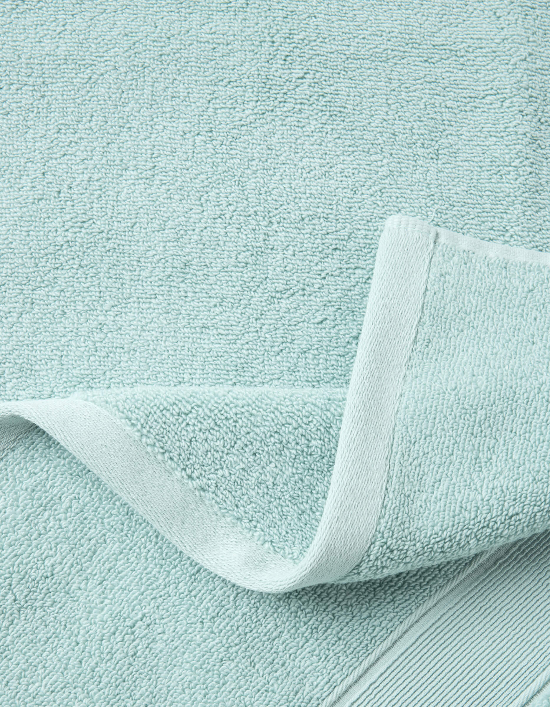 A close-up of the Luxe Bath Towel Set by Cozy Earth in light blue, featuring a woven border and folded corner that highlights the soft, absorbent fabric's texture.|Color:Tide