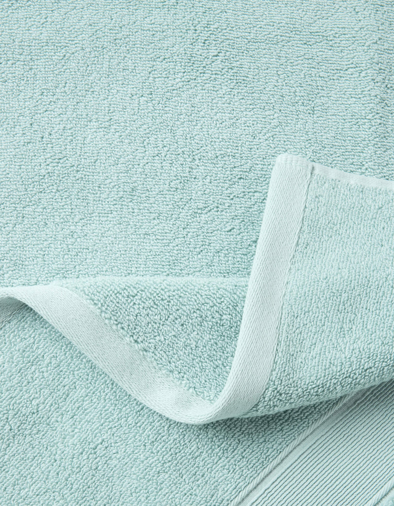 A close-up of the Luxe Bath Towel Set by Cozy Earth in light blue, featuring a woven border and folded corner that highlights the soft, absorbent fabric's texture.