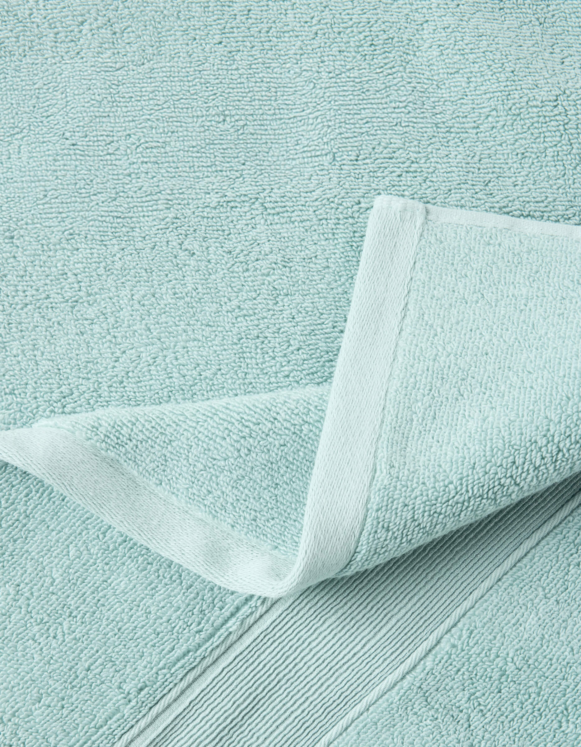 A close-up of a Luxe Bath Towel by Cozy Earth in light blue highlights its textured, soft, and absorbent fabric with detailed stitching and a neatly folded corner.|Color:Tide