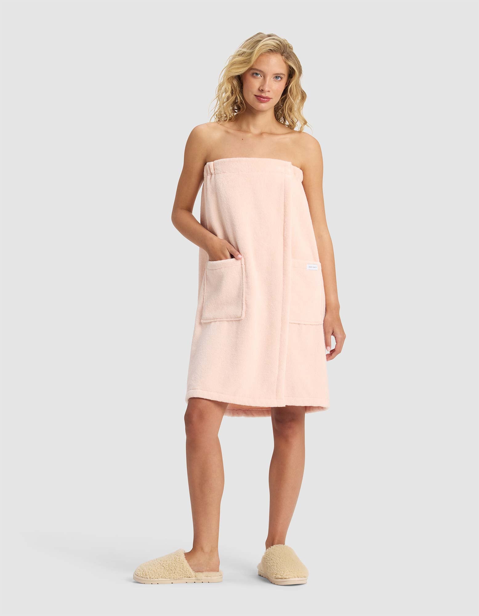 A woman with blonde hair wears the Cozy Earth Luxe Bath Wrap in light pink, featuring two front pockets, along with beige slippers, standing against a plain light gray background.