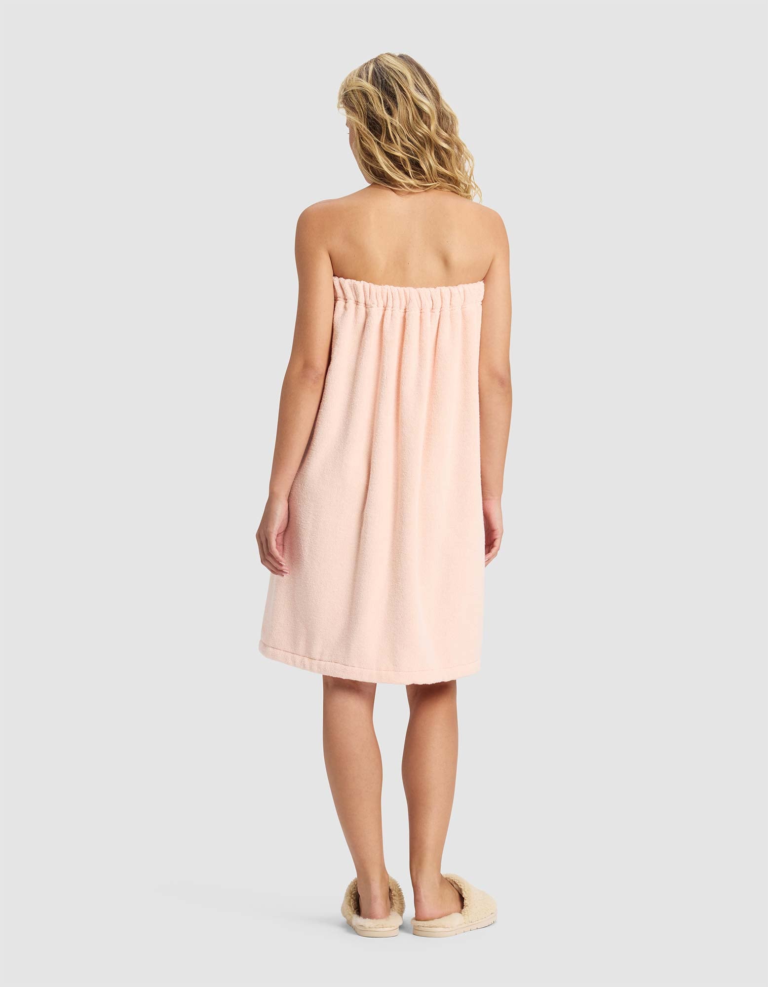 A woman with wavy blonde hair stands facing away, wearing the Cozy Earth Luxe Bath Wrap in soft light pink and beige slippers, set against a plain light gray background.
