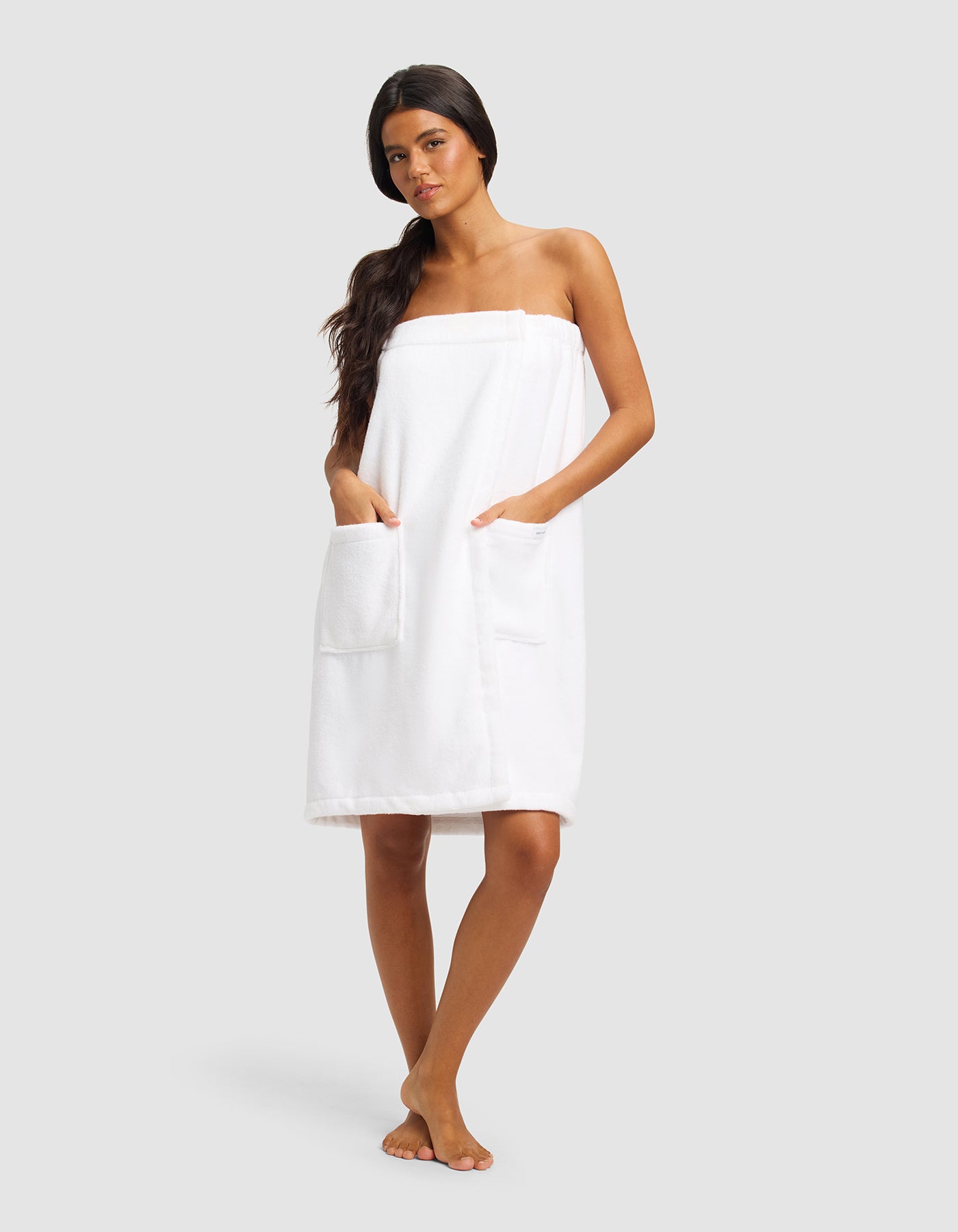 A woman with long dark hair stands barefoot against a plain background, wearing Cozy Earth's Luxe Bath Wrap featuring two large front pockets.