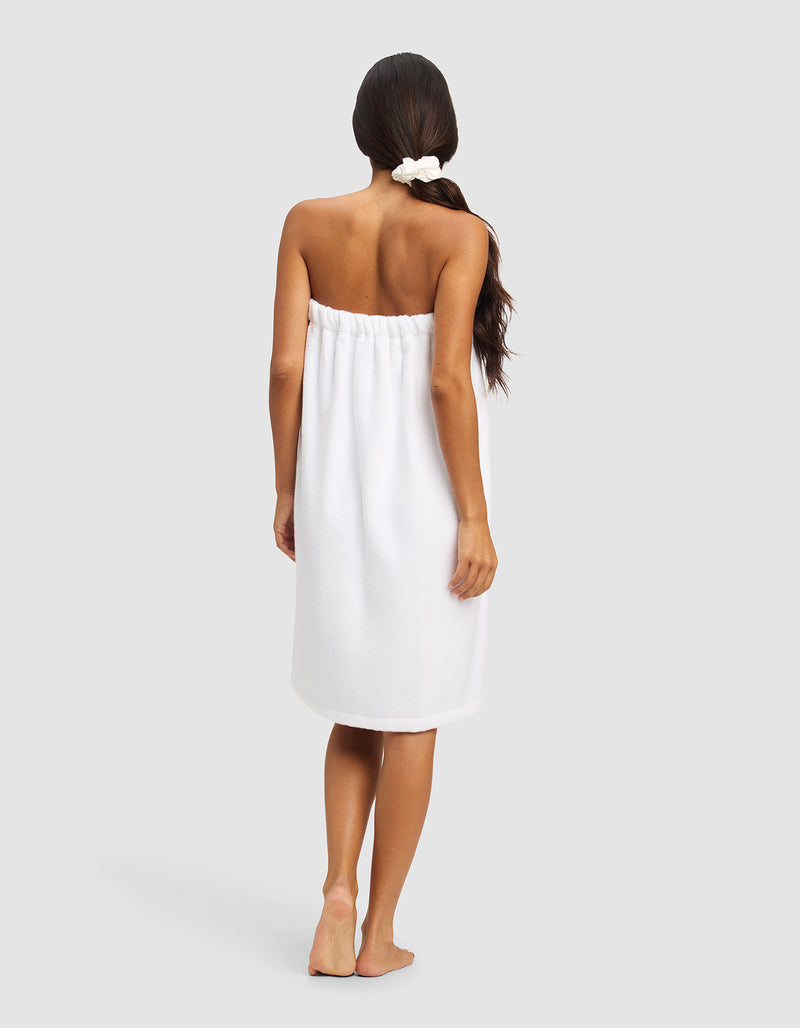 A woman with long dark hair in a low ponytail stands barefoot, facing away, wearing the Cozy Earth Luxe Bath Wrap against a plain light gray background.