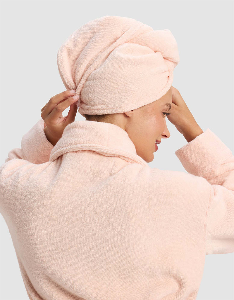 A person adjusts a Luxe Hair Towel by Cozy Earth, wrapped around their head, while wearing a light pink bathrobe, viewed from the back and side.