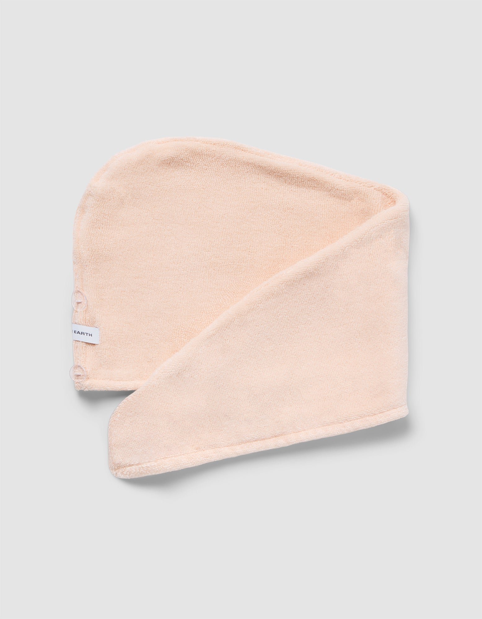 The Luxe Hair Towel by Cozy Earth is a soft, light beige microfiber towel with a white label and two snap buttons. Displayed partially folded on a light gray background, it highlights its rectangular shape and smooth texture.
