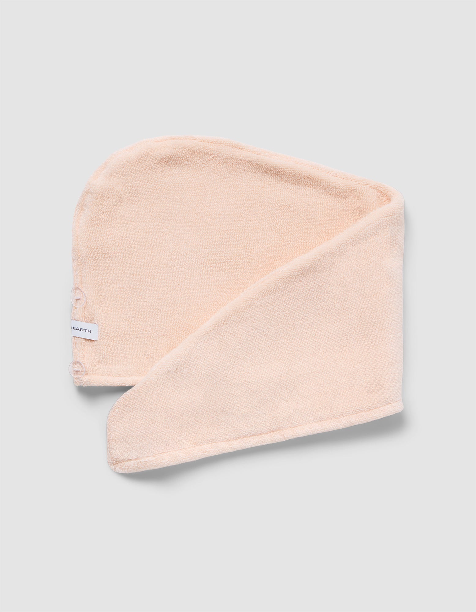 The Luxe Hair Towel by Cozy Earth is a soft, light beige microfiber towel with a white label and two snap buttons. Displayed partially folded on a light gray background, it highlights its rectangular shape and smooth texture.