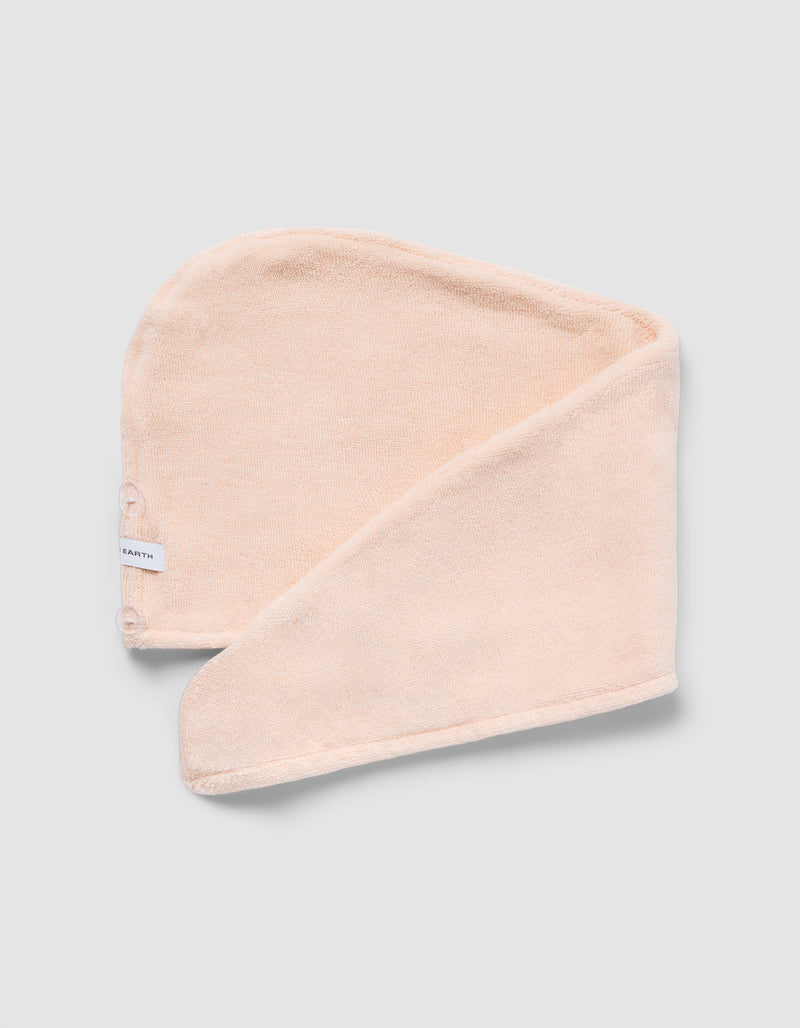 The Luxe Hair Towel by Cozy Earth is a soft, light beige microfiber towel with a white label and two snap buttons. Displayed partially folded on a light gray background, it highlights its rectangular shape and smooth texture.