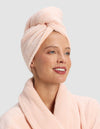 A smiling woman wears the Luxe Hair Towel by Cozy Earth and a light pink bathrobe, looking slightly to the side against a plain light gray background.|Color:Blossom