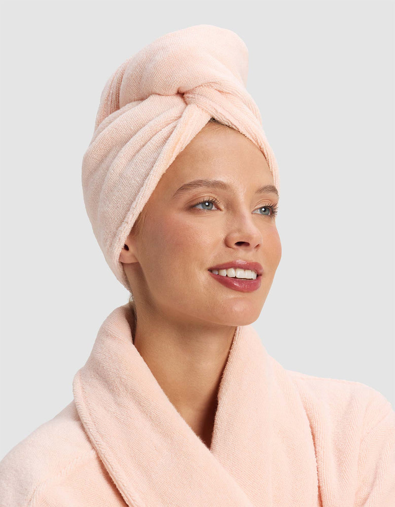 A smiling woman wears the Luxe Hair Towel by Cozy Earth and a light pink bathrobe, looking slightly to the side against a plain light gray background.
