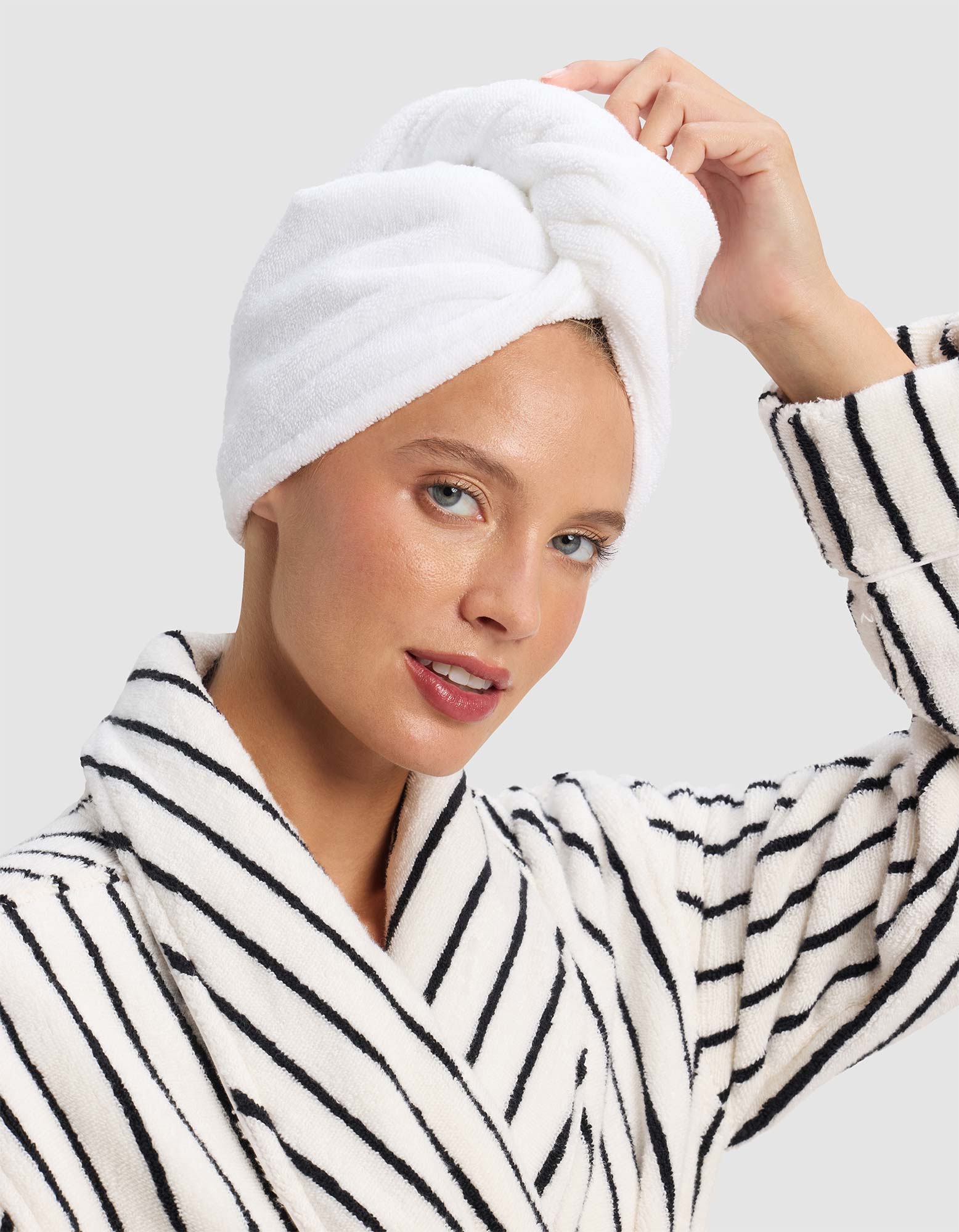 A woman smiles softly, touching the Cozy Earth Luxe Hair Towel wrapped around her head, while wearing a white bathrobe with black stripes against a plain light background. 