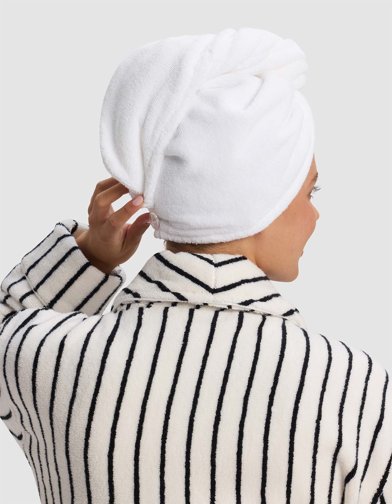 A person, seen from behind in a black and white striped robe, touches their head where a Cozy Earth Luxe Hair Towel is wrapped.