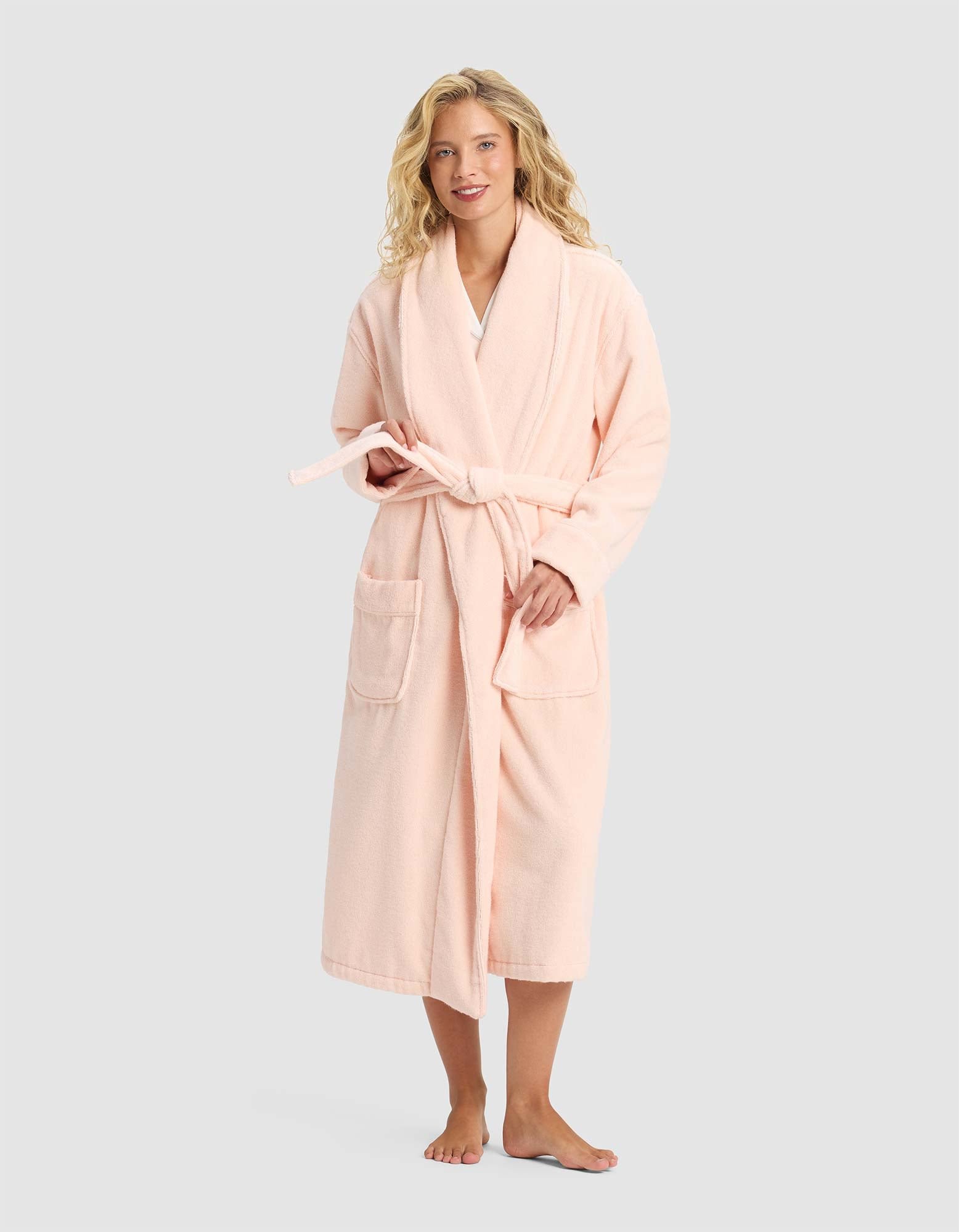 A smiling woman with blonde hair stands barefoot, facing forward in the Cozy Earth Luxe Bath Robe—a long, light pink robe with front pockets and a tie belt—set against a plain light gray background.|Color:Blossom