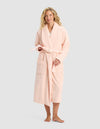 A smiling woman with blonde hair stands barefoot, facing forward in the Cozy Earth Luxe Bath Robe—a long, light pink robe with front pockets and a tie belt—set against a plain light gray background.|Color:Blossom
