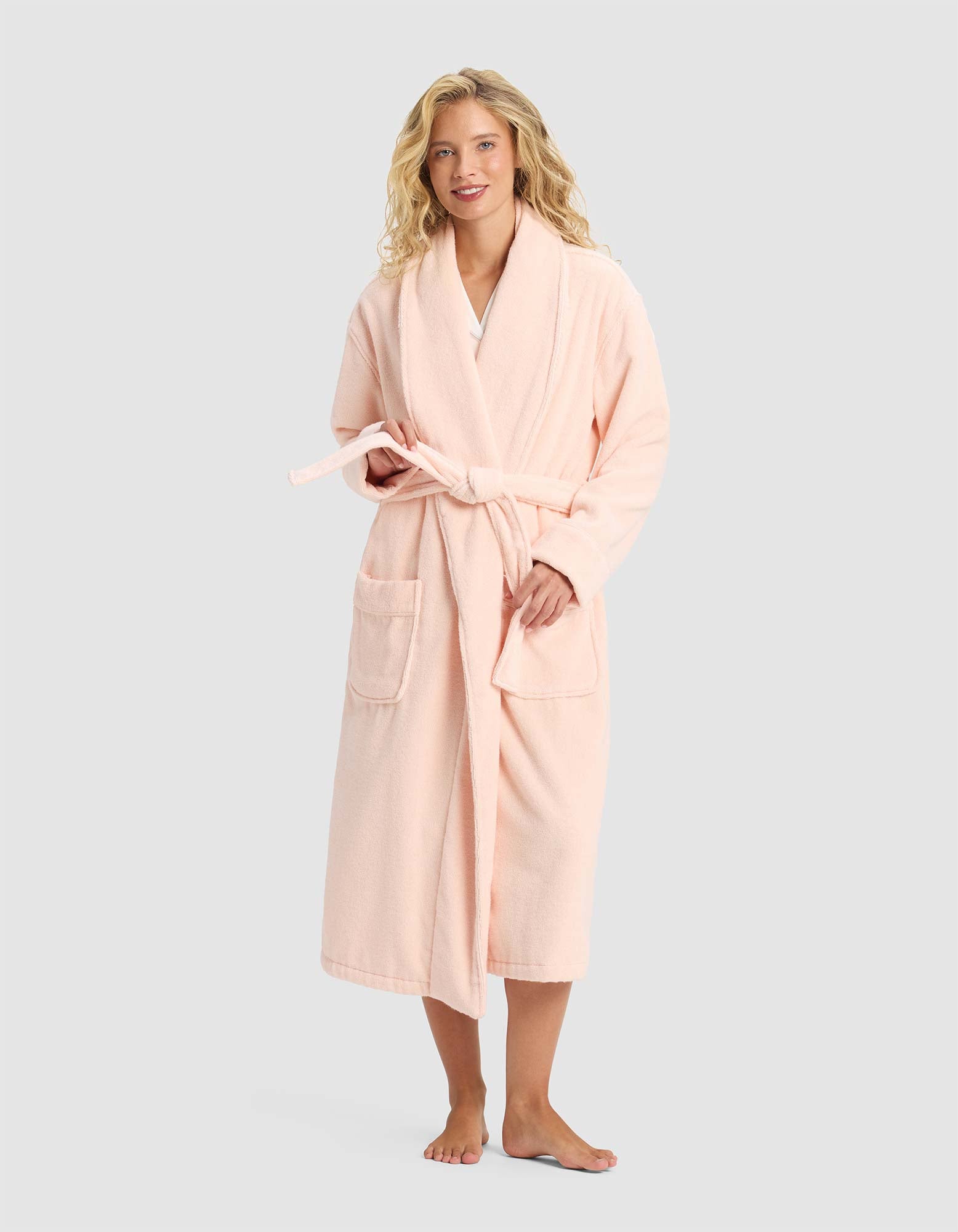 A smiling woman with blonde hair stands barefoot, facing forward in the Cozy Earth Luxe Bath Robe—a long, light pink robe with front pockets and a tie belt—set against a plain light gray background.|Color:Blossom