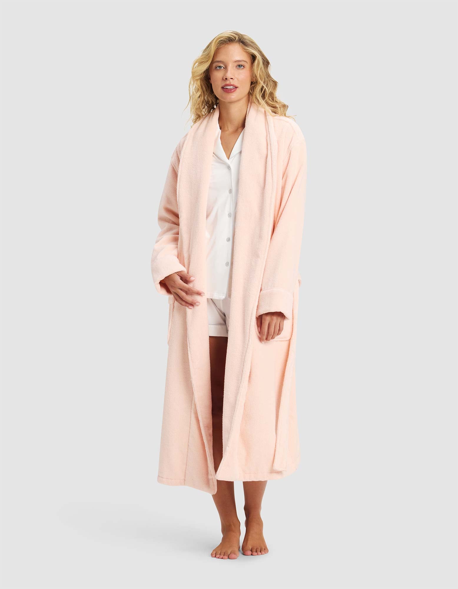 A woman stands barefoot against a plain light gray background, wearing Cozy Earth's Luxe Bath Robe in light pink over white pajama shorts and a matching button-up shirt.