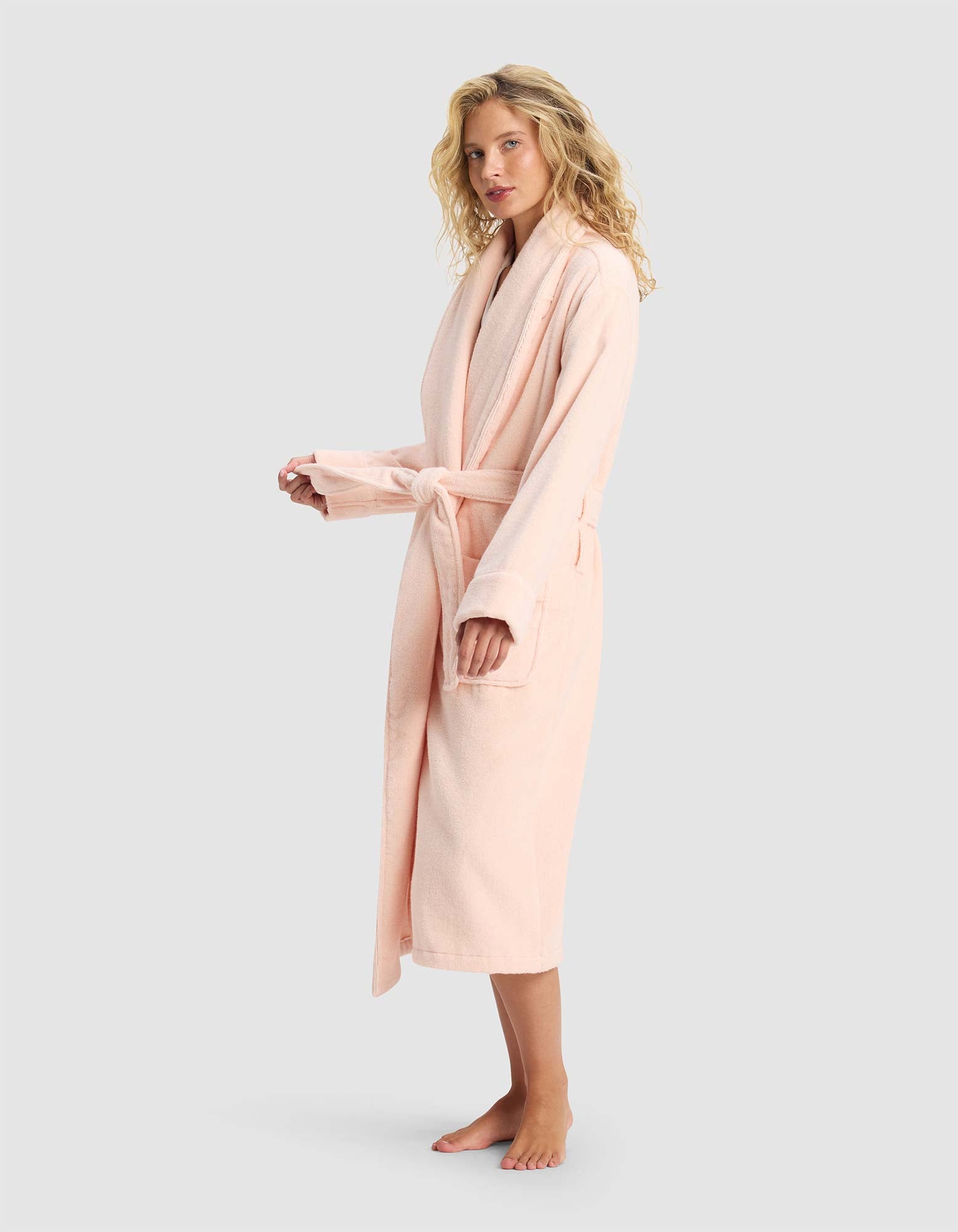 A woman with curly blonde hair wears the Cozy Earth Luxe Bath Robe in light pink, tied at the waist with two front pockets, standing barefoot against a plain light gray background.
