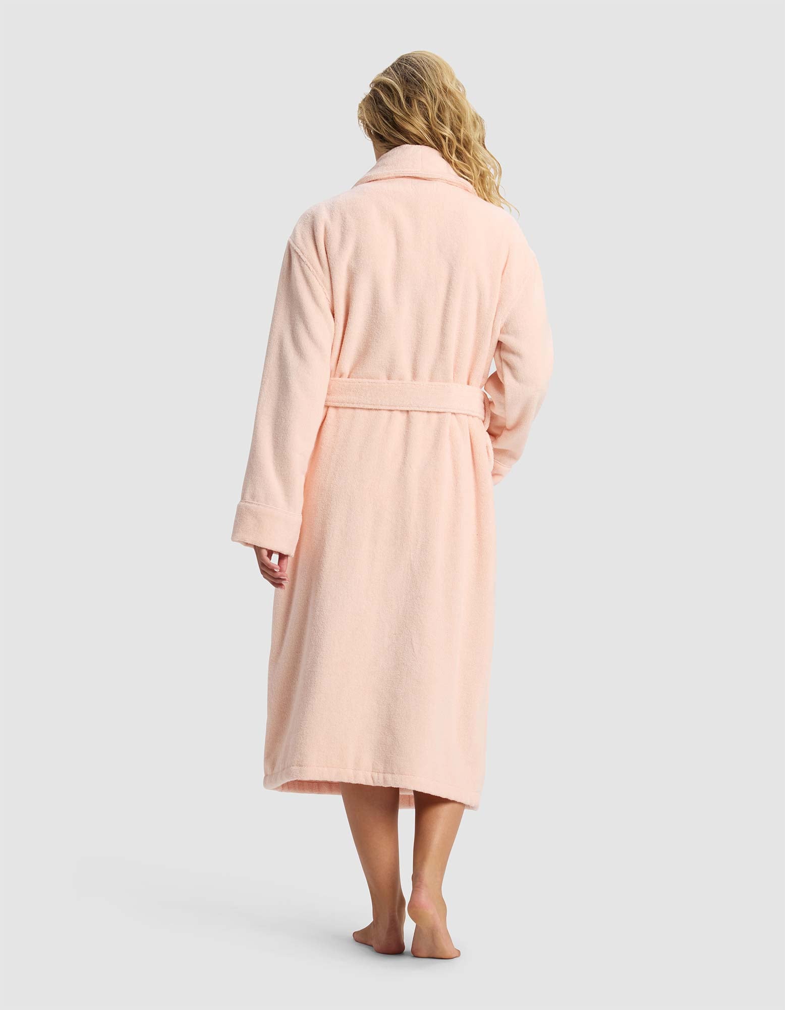 A person with long, wavy blonde hair stands barefoot, facing away, wearing the Cozy Earth Luxe Bath Robe in light pink with a belt against a plain light gray background.