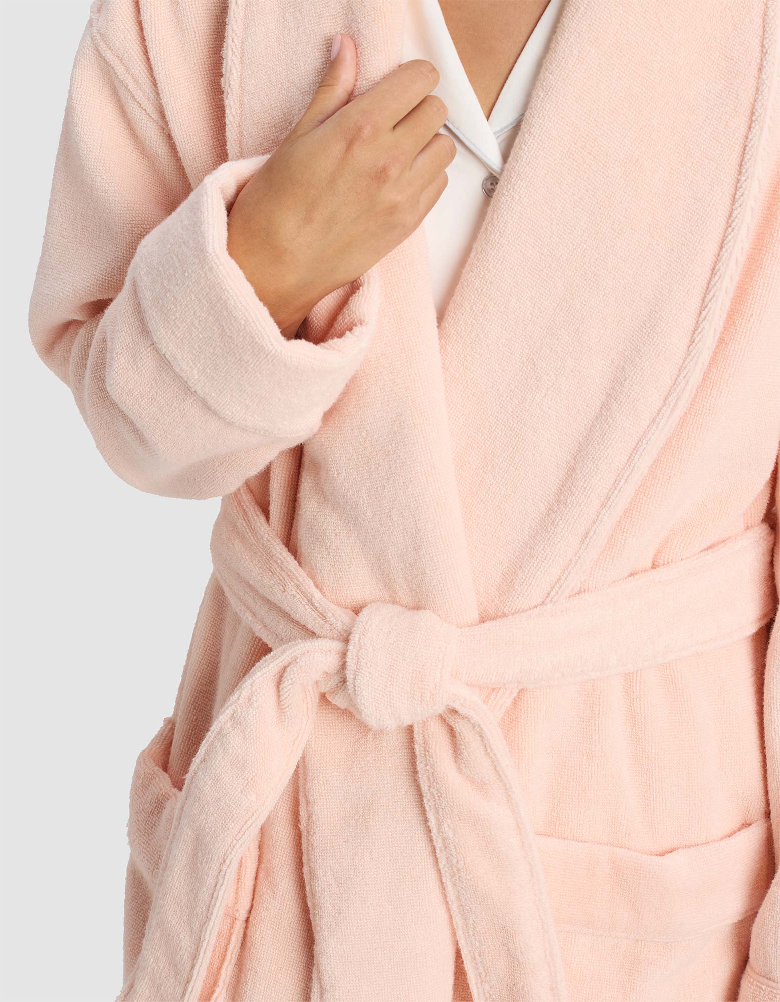 A person wears the Luxe Bath Robe by Cozy Earth in light pink, tied at the waist and holding the lapel. The background is plain and light-colored, with their face partially out of frame.