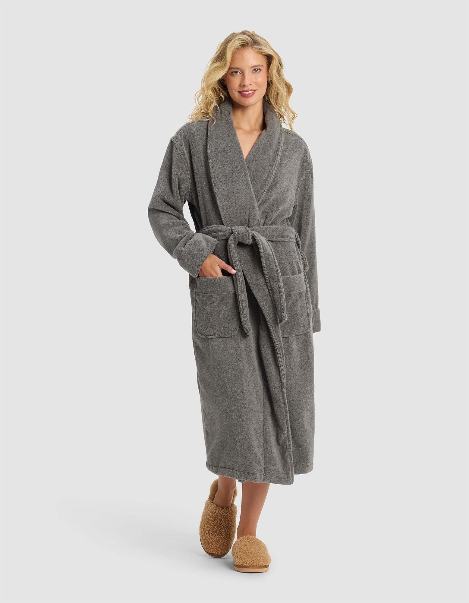 A woman with wavy blonde hair smiles while wearing the Cozy Earth Luxe Bath Robe in gray, featuring a belted waist and large pockets, paired with brown slippers, against a plain light background.