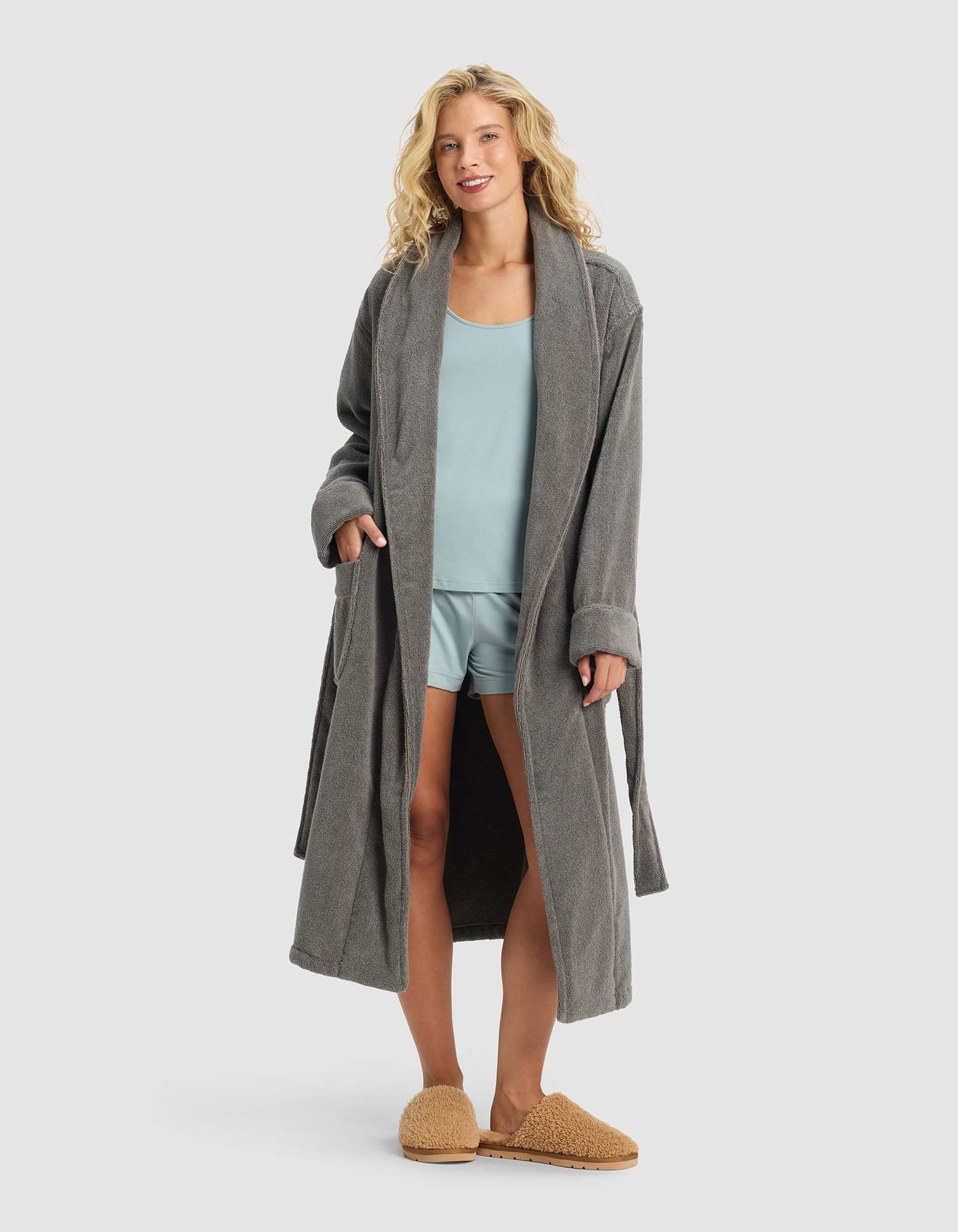 A woman smiles with her hands in the pockets of a Luxe Bath Robe by Cozy Earth, layering it over light blue pajamas and tan slippers against a plain light gray background.
