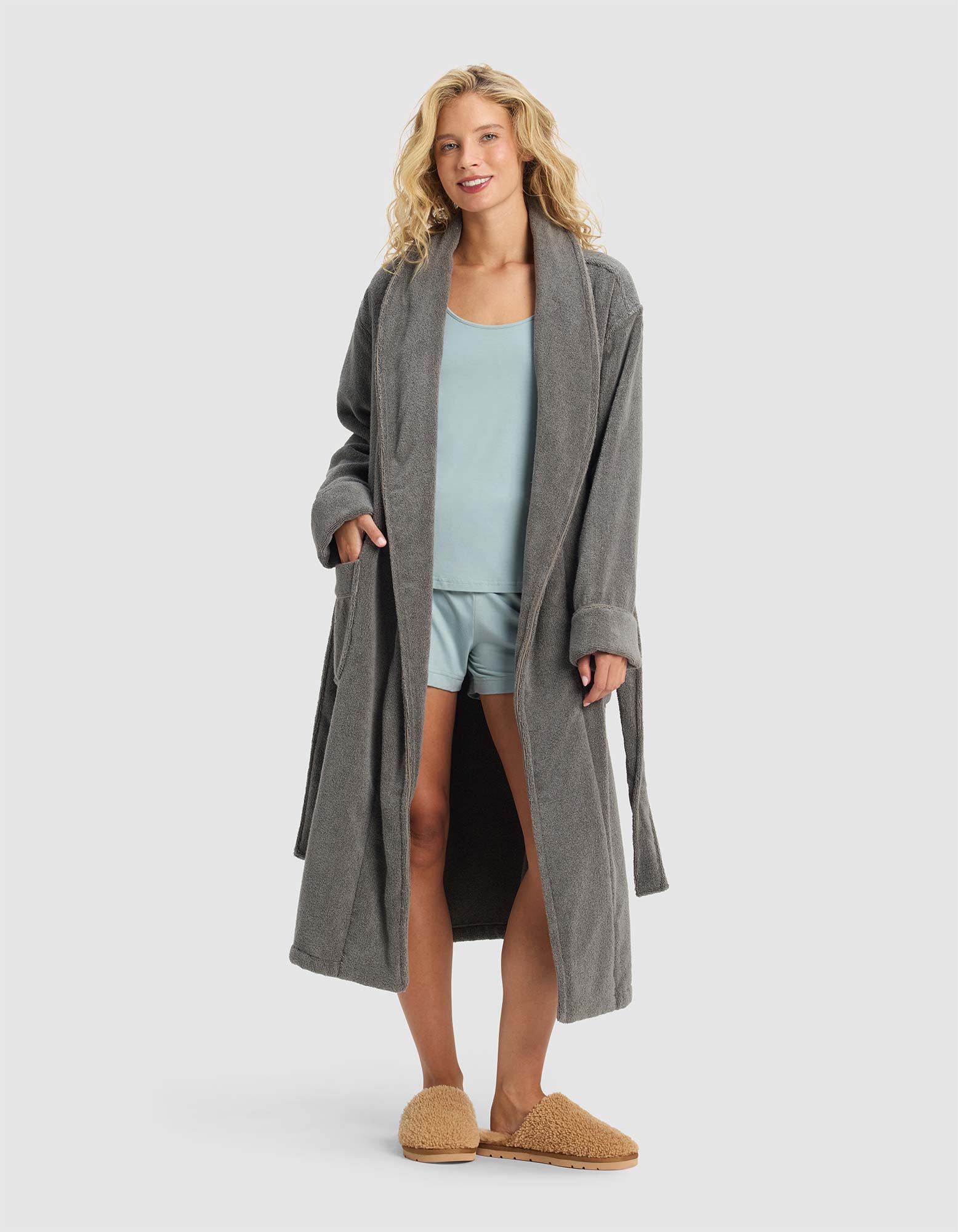A woman smiles with her hands in the pockets of a Luxe Bath Robe by Cozy Earth, layering it over light blue pajamas and tan slippers against a plain light gray background.|Color:Charcoal