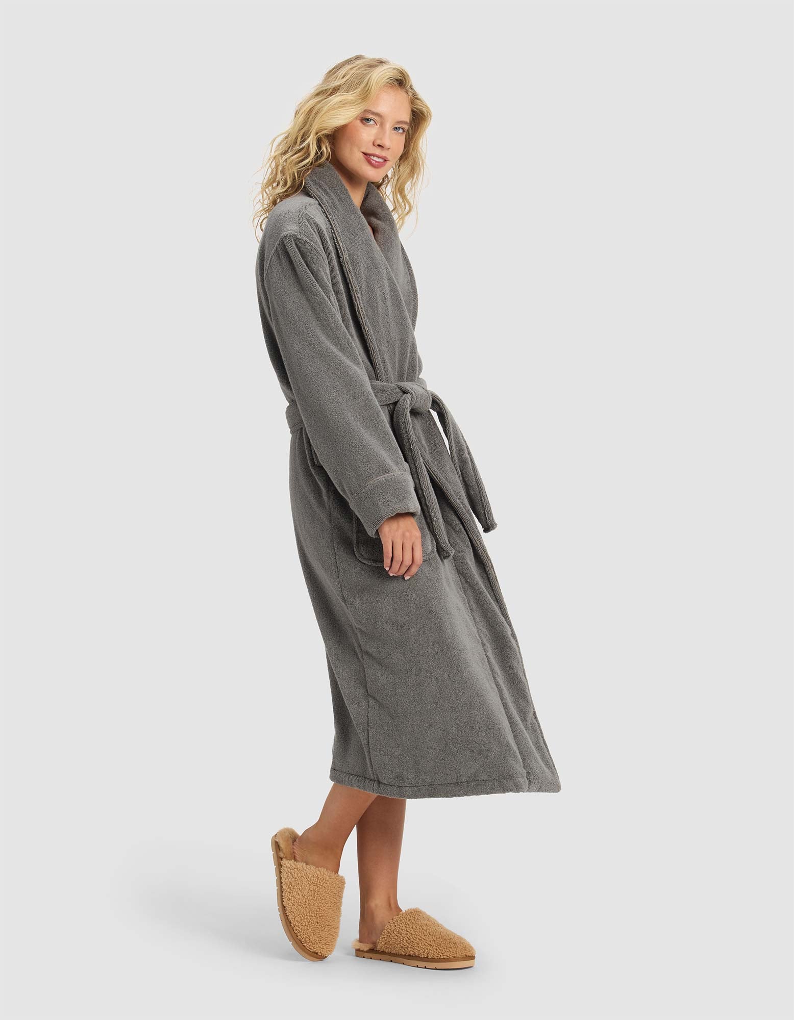 A person stands sideways in the Cozy Earth Luxe Bath Robe, a long gray robe with a waist belt, paired with tan fuzzy slippers against a plain light background.|Color:Charcoal