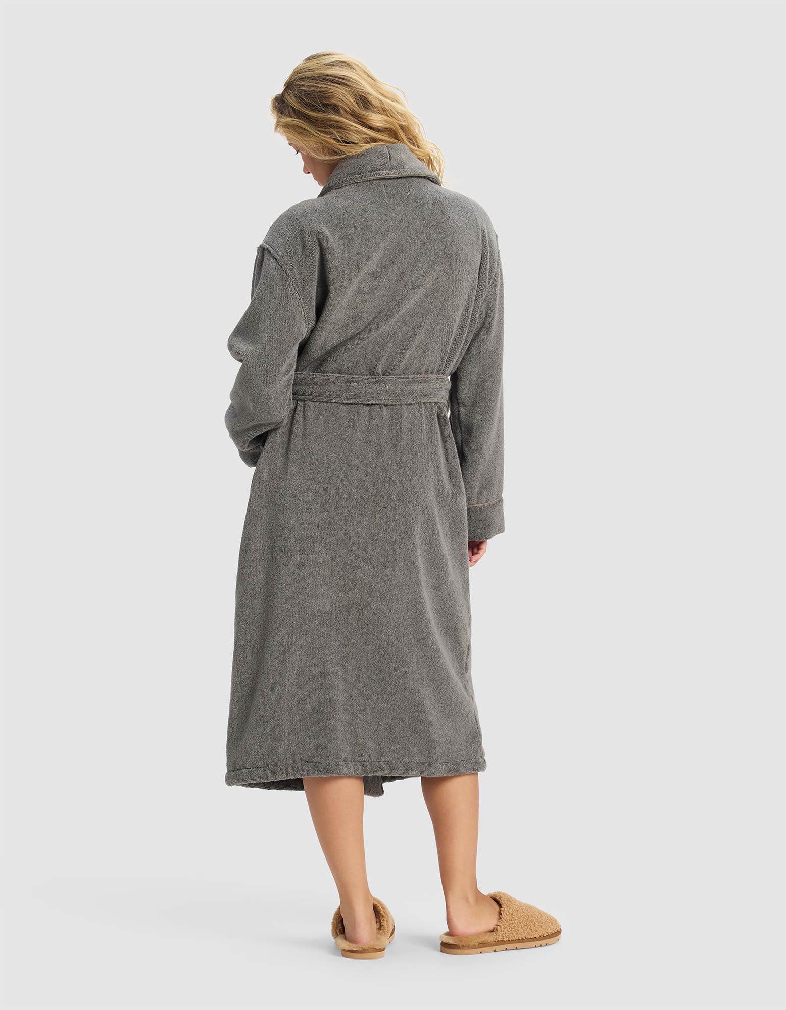 A woman with wavy blonde hair faces away, wearing the Luxe Bath Robe by Cozy Earth—a long gray robe with a belt—paired with tan fuzzy slippers, set against a plain light background.