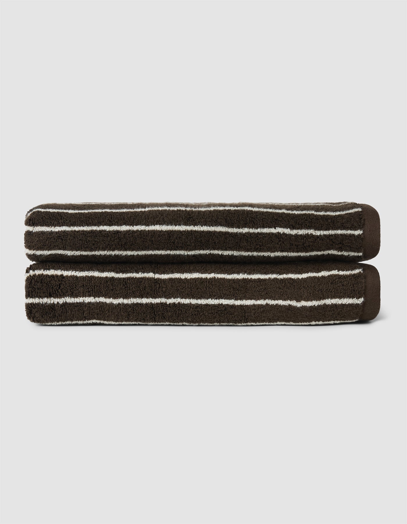 The Complete Striped Luxe Bath Bundle by Cozy Earth features two dark brown towels with thin, irregular white stripes, neatly rolled and stacked on a light gray background.|Color:Espresso Coconut Stripe