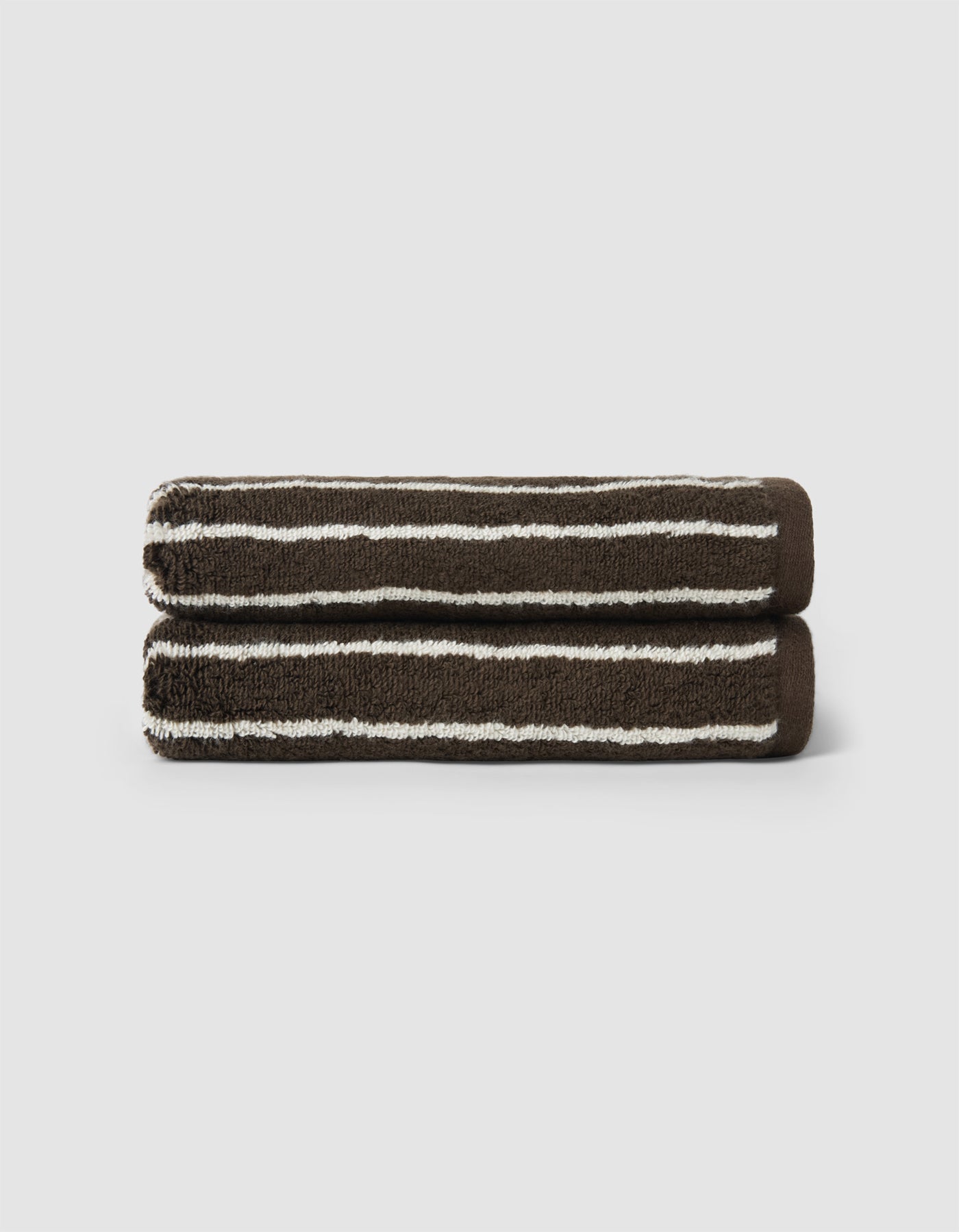 Two neatly rolled Striped Luxe Bath Towels by Cozy Earth, featuring dark brown with white horizontal stripes, are stacked atop each other against a plain light background.