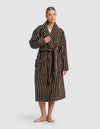 A woman stands barefoot in the Cozy Earth Striped Luxe Bath Robe—long, dark brown with white stripes, a shawl collar, belt, and large front pockets. She looks ahead, holding the lapel with one hand.|Color:Espresso Coconut Stripe