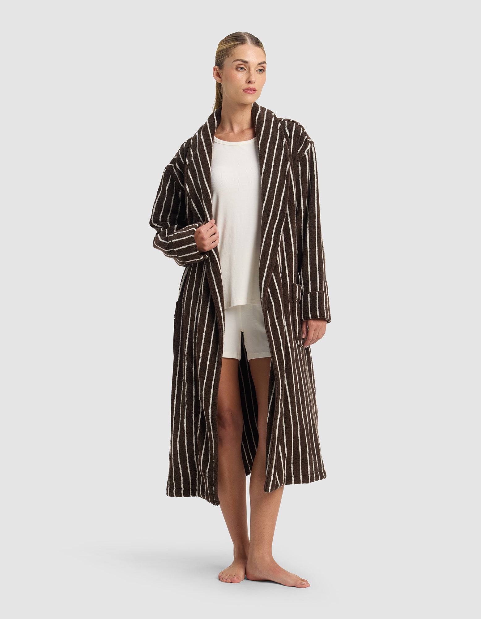 A woman stands barefoot against a light gray background, wearing the Cozy Earth Striped Luxe Bath Robe in brown with white vertical stripes, layered over a white t-shirt and white shorts.