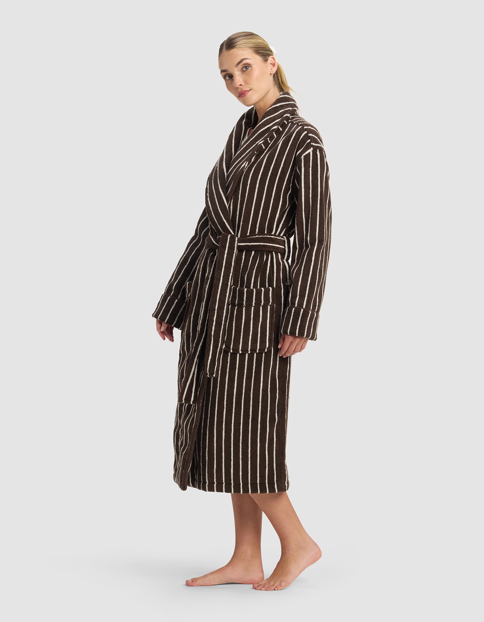A person stands barefoot in the Cozy Earth Striped Luxe Bath Robe, featuring brown fabric with white vertical stripes, large front pockets, and a belted waist, set against a light gray background.