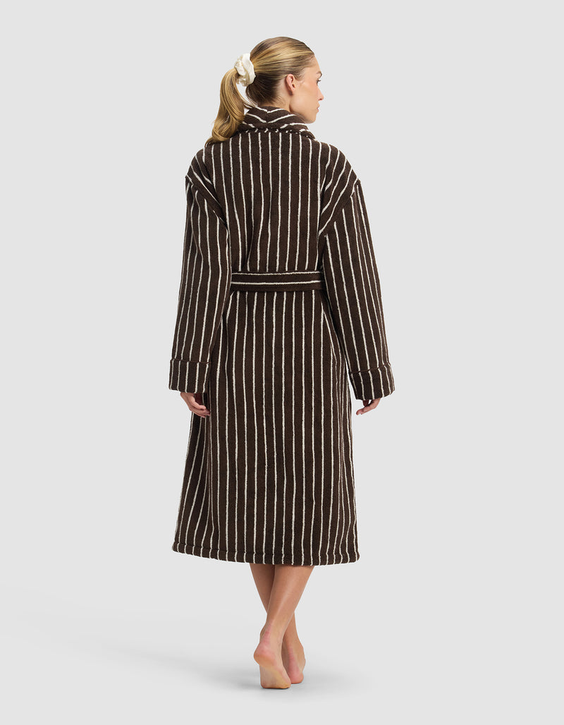 A person with light skin and blonde hair in a white scrunchie stands barefoot, back to camera, wearing the Cozy Earth Striped Luxe Bath Robe—a long, dark brown robe with vertical white stripes—against a plain light background.