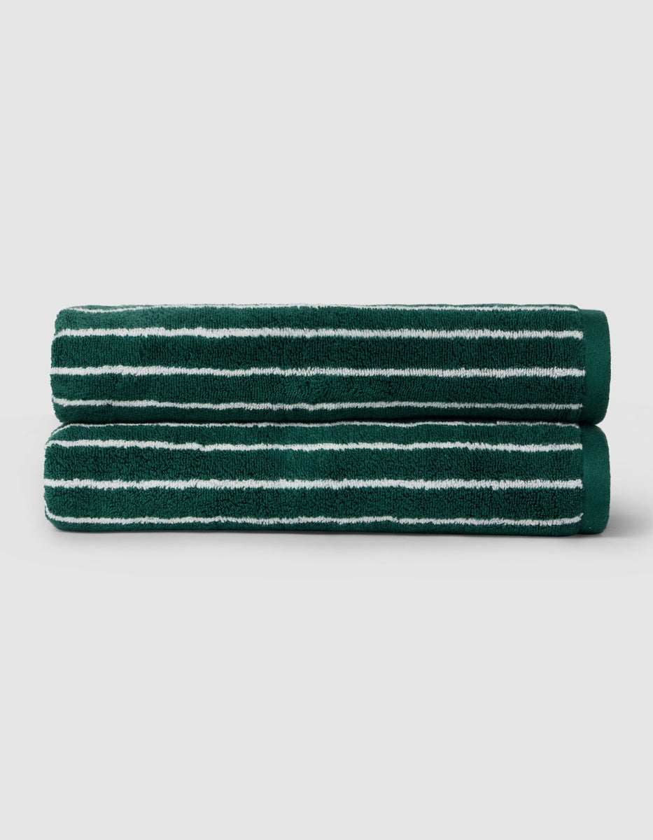 Two Luxe Bath Towels by Cozy Earth, dark green with white horizontal stripes, are neatly rolled and stacked on a light gray background.|Color:Uniform with Seashell Stripe