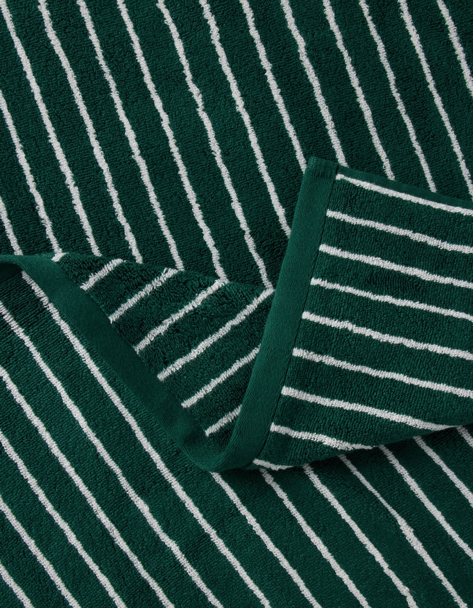 The Luxe Bath Towels by Cozy Earth in dark green with thin white stripes are neatly folded on a matching fabric background.