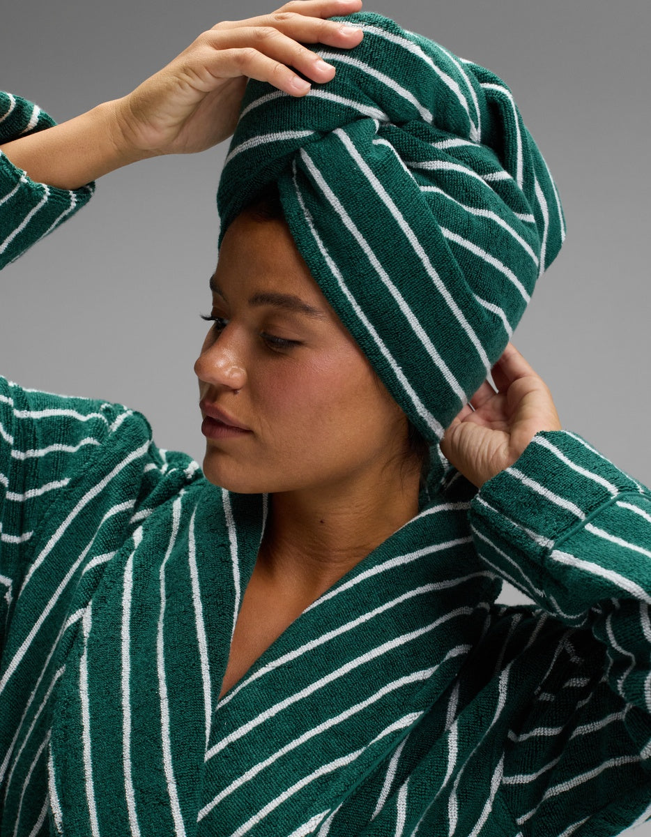 A woman wearing a green and white striped robe adjusts the Luxe Hair Towel by Cozy Earth on her head, looking to the side against a gray background.