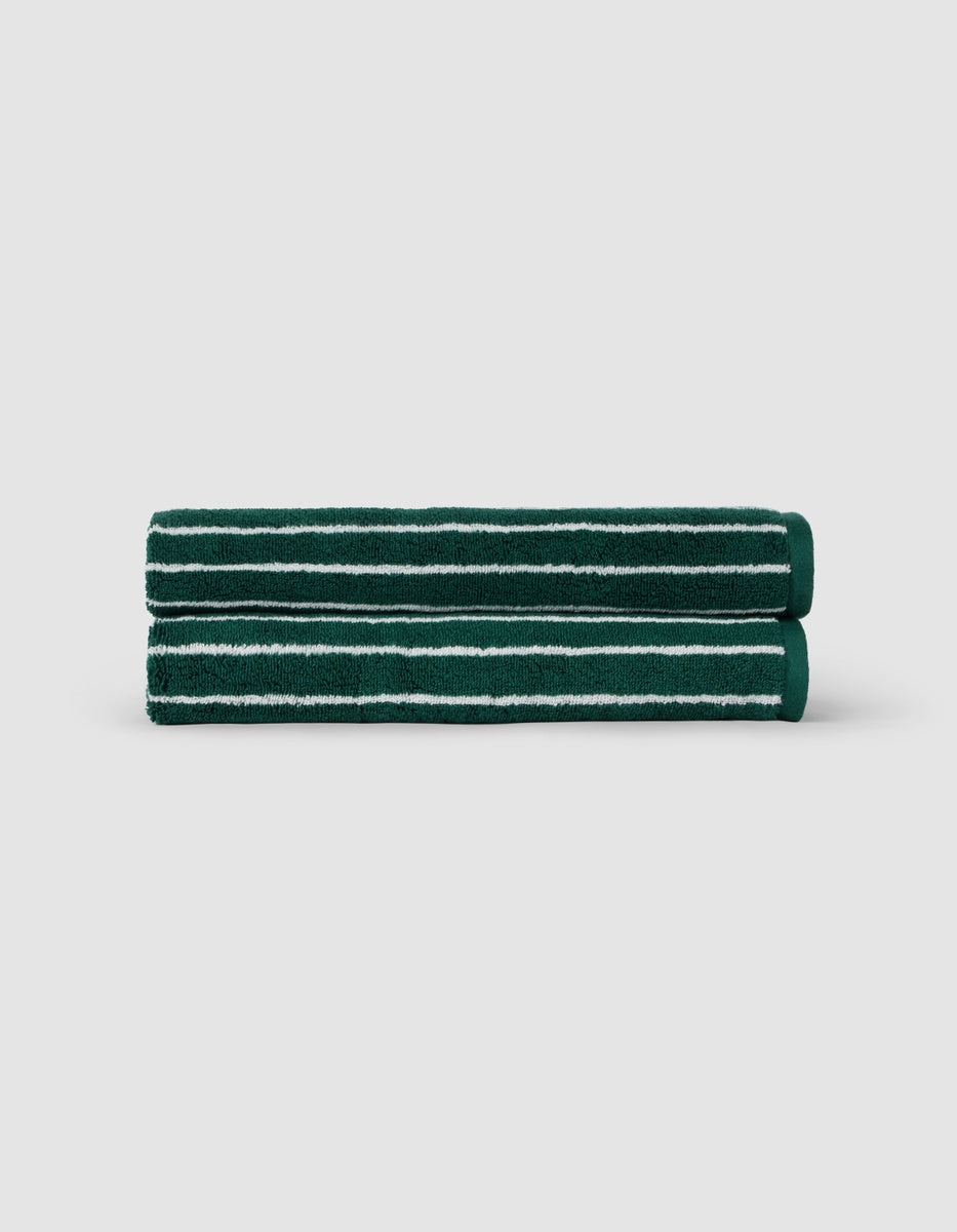 Two Luxe Hand Towels by Cozy Earth, dark green with white horizontal stripes, are neatly rolled and stacked against a plain light gray background.|Color:Uniform with Seashell Stripe