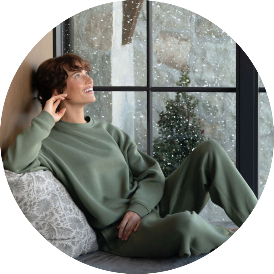 A woman wearing Cozy Earth CityScape Crewneck and Sweatpant set sits in front of a window as snow falls outside.