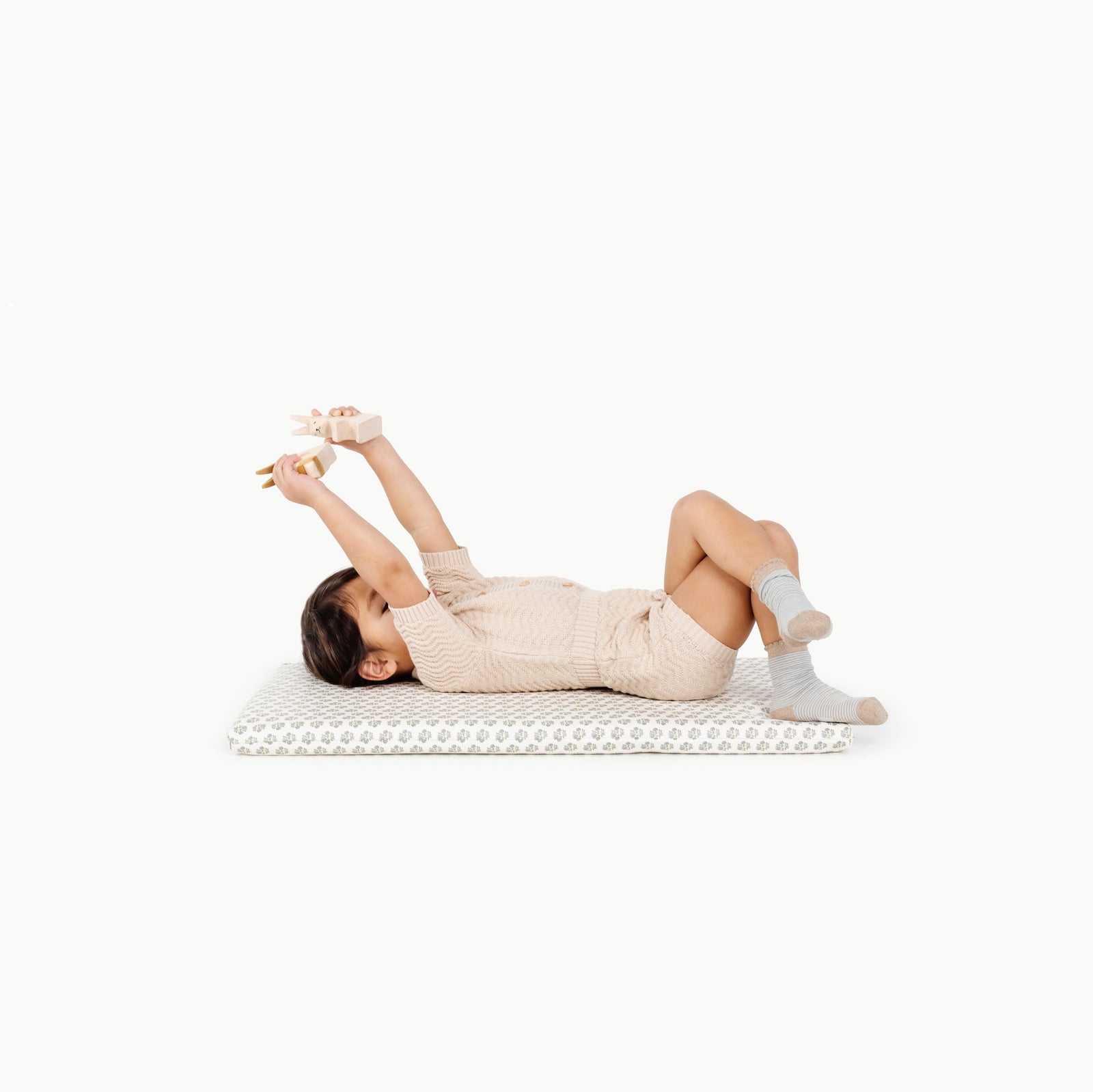 A child in a beige outfit and gray socks lies on a Gathre Wholesale Padded Changing Mat in Meadow, playing with wooden toys above their head against a white background.