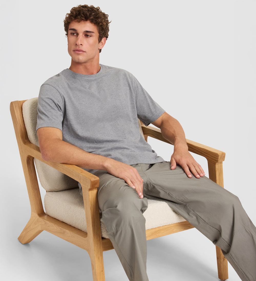 Man sitting on a wooden chair wearing a gray t-shirt and beige pants against a white background